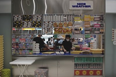 A small retail kiosk is filled with a variety of packaged food items like snacks, cereal boxes, and candy. Behind the counter, a person wearing a mask is organizing or checking inventory while standing in front of a display with a 'NICE WEATHER' sign above. The countertop has stacks of trays and a few other items.