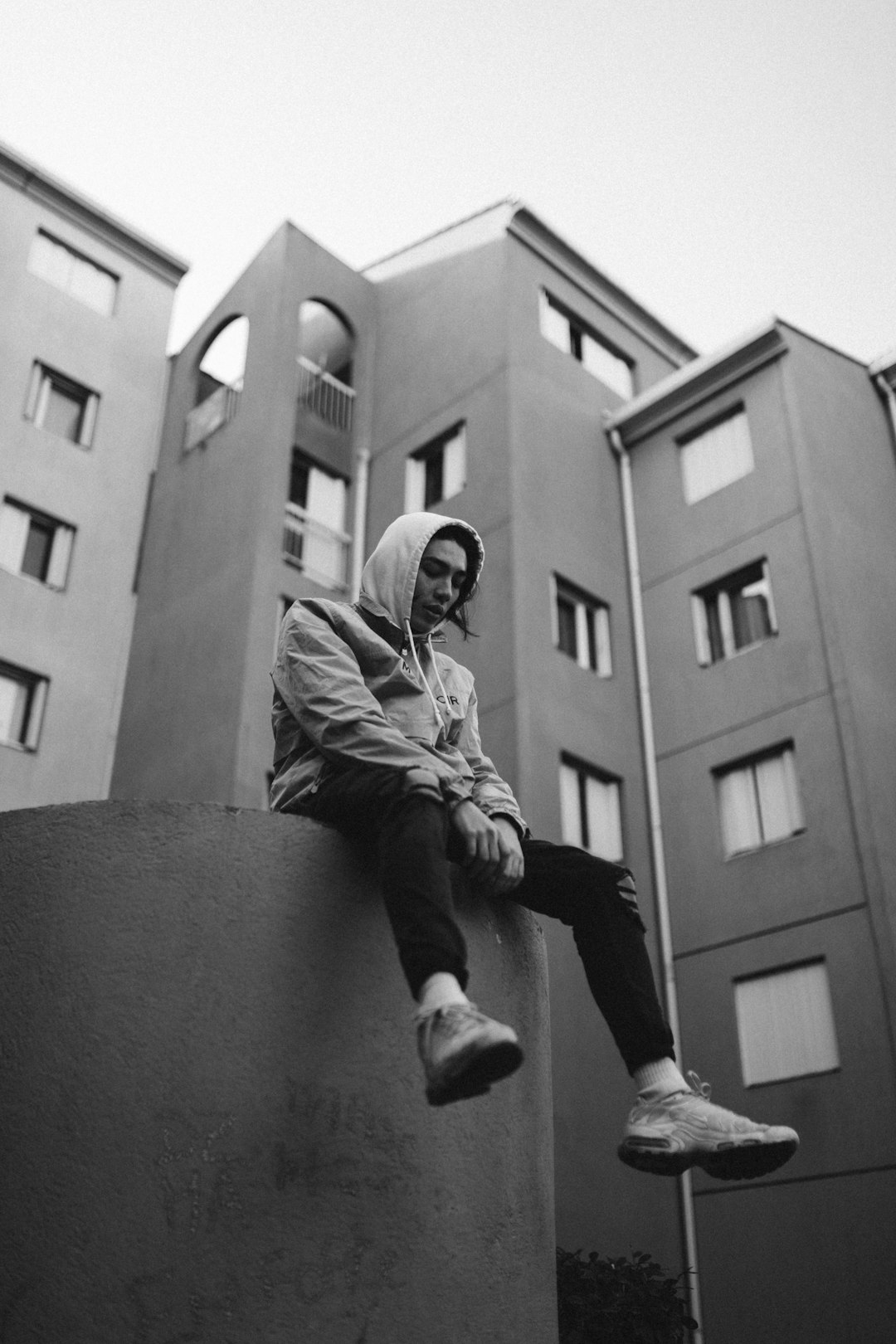 grayscale photo of man in jacket and pants sitting on concrete wall