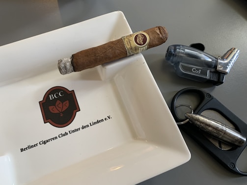 A cigar rests on a ceramic ashtray with the logo 'BCC' and the name 'Berliner Cigarrren Club Unter den Linden e.V.' inscribed on it. Nearby, there is a translucent, dark-colored lighter and a metal cigar cutter featuring a bullet-like design.