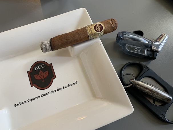A cigar rests on a ceramic ashtray with the logo 'BCC' and the name 'Berliner Cigarrren Club Unter den Linden e.V.' inscribed on it. Nearby, there is a translucent, dark-colored lighter and a metal cigar cutter featuring a bullet-like design.