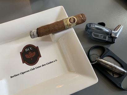 A cigar rests on a ceramic ashtray with the logo 'BCC' and the name 'Berliner Cigarrren Club Unter den Linden e.V.' inscribed on it. Nearby, there is a translucent, dark-colored lighter and a metal cigar cutter featuring a bullet-like design.
