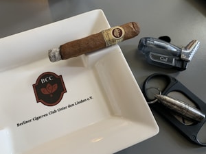 A cigar rests on a ceramic ashtray with the logo 'BCC' and the name 'Berliner Cigarrren Club Unter den Linden e.V.' inscribed on it. Nearby, there is a translucent, dark-colored lighter and a metal cigar cutter featuring a bullet-like design.