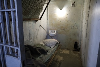 A small, dimly lit prison cell with a worn, metal bed against one wall, topped with a thin mattress and pillow. Chains hold up the bed, and a small sink is mounted on the opposite wall. The walls are concrete with signs of wear and chipping, and a single light bulb provides sparse illumination.
