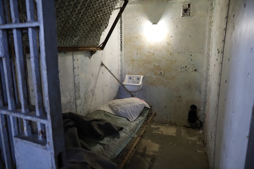 A small, dimly lit prison cell with a worn, metal bed against one wall, topped with a thin mattress and pillow. Chains hold up the bed, and a small sink is mounted on the opposite wall. The walls are concrete with signs of wear and chipping, and a single light bulb provides sparse illumination.