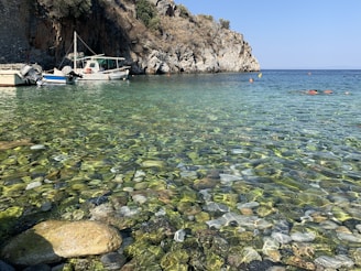 A tranquil coastal scene features clear, shallow waters with smooth, multicolored pebbles visible beneath the surface. Several boats, including a fishing boat, are moored near a rocky shoreline with a rugged cliff. Sparse vegetation grows atop the cliff under a clear blue sky.