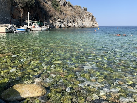 A tranquil coastal scene features clear, shallow waters with smooth, multicolored pebbles visible beneath the surface. Several boats, including a fishing boat, are moored near a rocky shoreline with a rugged cliff. Sparse vegetation grows atop the cliff under a clear blue sky.