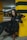 A person with long dark hair crouches next to a large Doberman dog in an underground parking garage. The person is dressed in black leather, matching the sleek coat of the dog. They both stand beside a yellow and black striped pillar, giving a contrasting and bold appearance.