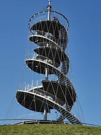 A tall spiral observation tower stands against a clear blue sky, featuring multiple circular platforms connected by spiral staircases. The structure is supported by a network of cables. The base is surrounded by a grassy hill.