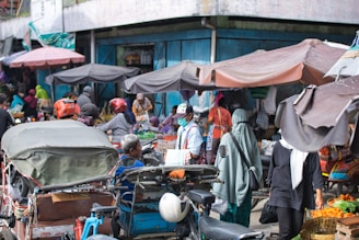 A bustling street market scene filled with people wearing various clothing styles, including helmets and headscarves. Numerous stalls are covered with tarps, offering a variety of goods. A motorbike with a sidecar is prominently featured, surrounded by shoppers and vendors.