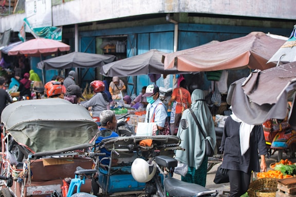 A bustling street market scene filled with people wearing various clothing styles, including helmets and headscarves. Numerous stalls are covered with tarps, offering a variety of goods. A motorbike with a sidecar is prominently featured, surrounded by shoppers and vendors.