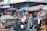 A bustling street market scene filled with people wearing various clothing styles, including helmets and headscarves. Numerous stalls are covered with tarps, offering a variety of goods. A motorbike with a sidecar is prominently featured, surrounded by shoppers and vendors.