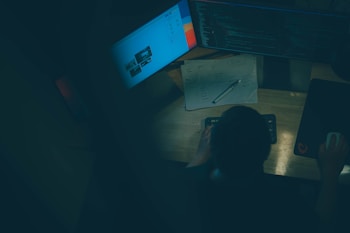 A person is seated at a wooden desk, illuminated by the glow of computer screens. There are two monitors displaying code and webpage layouts. Papers with sketches and a pen rest on the desk, and the individual appears to be working with a mouse and keyboard.