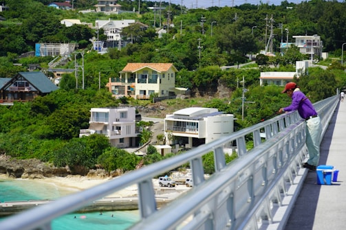 A person wearing a purple shirt and red cap is fishing from a bridge that overlooks a coastal area. Several modern houses are nestled among lush green trees. Below, a sandy beach merges with clear blue water. Power lines and a few parked vehicles are seen in the distance.