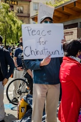 A person holds a sign that reads 'Peace for Chinatown!' during a public demonstration. Several people are present, both in the foreground and background, some wearing masks. A bicycle is visible, and you're surrounded by trees and buildings, indicating an urban setting.
