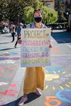 A person wearing a face mask holds a sign that reads 'Protect Our Mothers Fathers Grandmas Grandpas' in both English and Chinese characters. The background features a sunny day in a park, with colorful chalk drawings on the pavement and other people walking around.