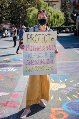 A person wearing a face mask holds a sign that reads 'Protect Our Mothers Fathers Grandmas Grandpas' in both English and Chinese characters. The background features a sunny day in a park, with colorful chalk drawings on the pavement and other people walking around.