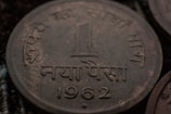 A close-up view of an old coin with Hindi script and the year 1962 etched on it. The coin appears to have some tarnish, indicating its age. The denomination is one 'Naya Paisa'.