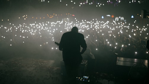 A musician is silhouetted against the glow of a crowd holding up lights, likely from phones, creating a sea of small bright dots. The atmosphere appears to be energetic and immersive, suggesting a live concert or music performance.