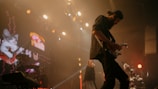 A guitarist is performing on stage, bathed in warm lighting. A digital screen in the background displays animated images, including a cat playing a guitar. The atmosphere appears lively with several spotlights creating dynamic lighting effects.