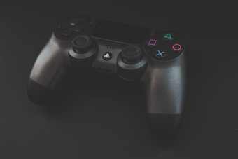 A black video game controller with colored buttons is positioned against a dark background. The buttons include triangles, squares, circles, and crosses highlighted in bright colors such as green, red, pink, and blue.