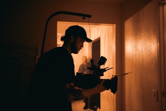 A person wearing a baseball cap operates a professional video camera in a dimly lit hallway. The walls are bathed in warm, amber lighting. In the background, another person is faintly visible down the hallway.