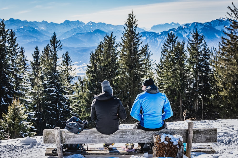 man in blue jacket sitting on brown wooden bench looking at the mountains during daytime