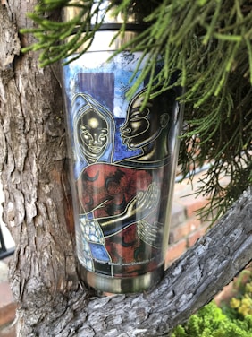 A travel mug nestled within the branches of a tree. The mug features an artistic design depicting two stylized figures with intricate facial details and elongated hands, set against a vibrant background. The artwork displays a combination of bold red and blue colors.