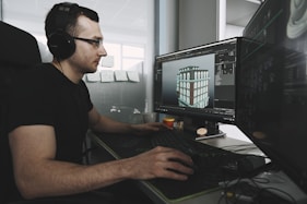 A person wearing black headphones is sitting at a desk and working on two computer monitors. The screens display 3D modeling software showing a building model. Sticky notes are on a glass partition in the background. The workspace includes a keyboard, a mouse, and a matte black desk mat.