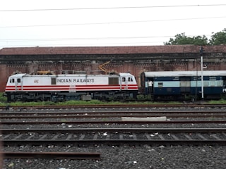 A train engine and luggage coach from Indian Railways are standing on a railway track. The engine is white with red stripes and labeled with 'Indian Railways'. In the background, there is an old brick building with a tiled roof. The area is surrounded by greenery.