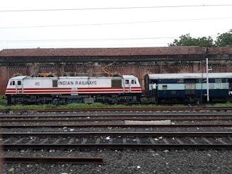 A train engine and luggage coach from Indian Railways are standing on a railway track. The engine is white with red stripes and labeled with 'Indian Railways'. In the background, there is an old brick building with a tiled roof. The area is surrounded by greenery.