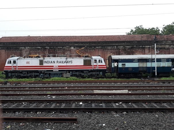 A train engine and luggage coach from Indian Railways are standing on a railway track. The engine is white with red stripes and labeled with 'Indian Railways'. In the background, there is an old brick building with a tiled roof. The area is surrounded by greenery.