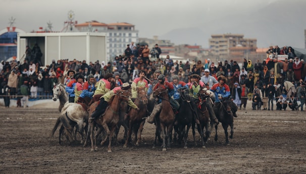 A group of horseback riders engaged in a competitive activity on a muddy field surrounded by a large crowd of spectators. The riders are dressed in colorful outfits, and the audience appears captivated by the event. The background features buildings under a cloudy, gray sky.