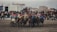 A group of horseback riders engaged in a competitive activity on a muddy field surrounded by a large crowd of spectators. The riders are dressed in colorful outfits, and the audience appears captivated by the event. The background features buildings under a cloudy, gray sky.