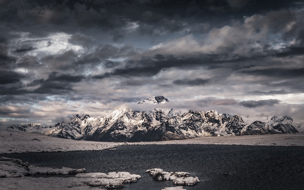 snow covered mountain near body of water under cloudy sky