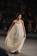 A woman walks confidently down a runway wearing an elegant, long, cream-colored dress adorned with intricate lace patterns. Her hair is styled in an updo, and she wears statement earrings. Seated spectators in the background watch attentively, creating a focused ambiance.