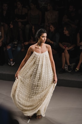 A woman walks confidently down a runway wearing an elegant, long, cream-colored dress adorned with intricate lace patterns. Her hair is styled in an updo, and she wears statement earrings. Seated spectators in the background watch attentively, creating a focused ambiance.