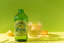 A green glass bottle with a label that reads 'Bundaberg Lemon, Lime & Bitters' situated on a yellow surface with a vibrant green background. Nearby, there is a clear glass containing a beverage with a lemon slice and a lemon wedge placed on the surface.