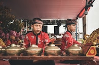 A group of young musicians dressed in traditional attire are playing musical instruments. They are wearing red and black clothing with intricate patterns and headgear. The setting appears to be a stage, with microphones visible, suggesting a performance. The instruments being played are gamelans, traditional Indonesian percussion instruments, made of metallic materials.