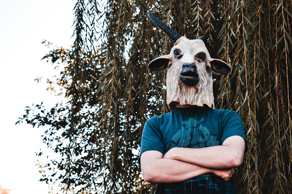 person in blue shirt wearing white and black animal skull mask standing near brown tree during