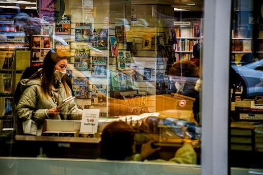 A woman wearing a mask is browsing books inside a bookstore, surrounded by shelves filled with various art books. Reflections from the window create an overlapping view of the interior and the street. The setting is warm and cozy with a mix of modern and classical elements.