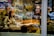 A woman wearing a mask is browsing books inside a bookstore, surrounded by shelves filled with various art books. Reflections from the window create an overlapping view of the interior and the street. The setting is warm and cozy with a mix of modern and classical elements.