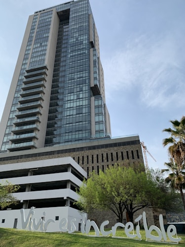 A modern high-rise building with a glass facade rises into the sky, with distinctive balconies on the left side. In the foreground, there are trees and greenery, along with a white sign reading 