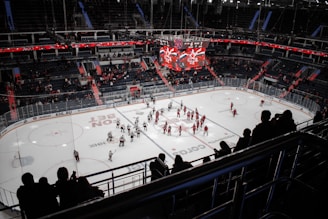 An indoor ice hockey stadium with players on the rink shaking hands near the center. Spectators are visible in the stands, some standing and some seated. The arena is well-lit with a large overhead scoreboard displaying scores and team logos. The dominant colors of team jerseys are red and white.