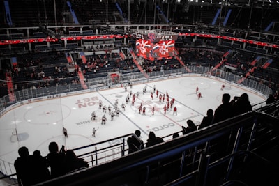 An indoor ice hockey stadium with players on the rink shaking hands near the center. Spectators are visible in the stands, some standing and some seated. The arena is well-lit with a large overhead scoreboard displaying scores and team logos. The dominant colors of team jerseys are red and white.