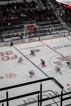 An ice hockey game taking place in an indoor arena with players wearing red and white uniforms on the ice. The rink is surrounded by glass panels, and there are spectators seated in the stands. A referee is visible on the ice, and advertisements line the perimeter of the rink.