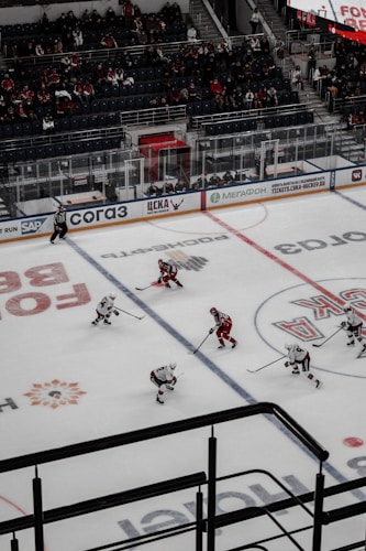 An ice hockey game taking place in an indoor arena with players wearing red and white uniforms on the ice. The rink is surrounded by glass panels, and there are spectators seated in the stands. A referee is visible on the ice, and advertisements line the perimeter of the rink.