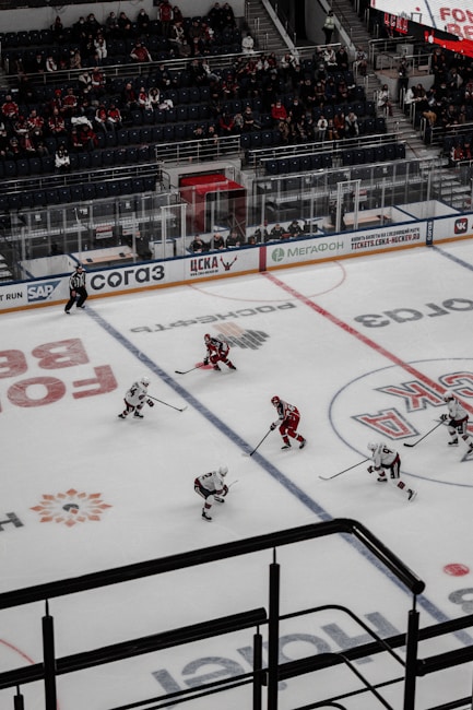 An ice hockey game taking place in an indoor arena with players wearing red and white uniforms on the ice. The rink is surrounded by glass panels, and there are spectators seated in the stands. A referee is visible on the ice, and advertisements line the perimeter of the rink.