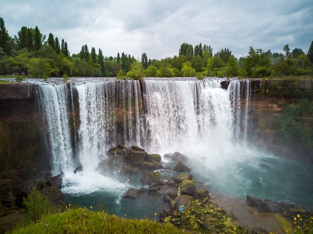 waterfalls near green trees under white clouds during daytime