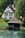 A large, traditional Swiss-style house with a steep roof and multiple windows is surrounded by lush greenery. In the foreground, there is a wooden boathouse next to calm water. A Swiss flag is visible near the boathouse, indicating the location.