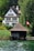 A large, traditional Swiss-style house with a steep roof and multiple windows is surrounded by lush greenery. In the foreground, there is a wooden boathouse next to calm water. A Swiss flag is visible near the boathouse, indicating the location.
